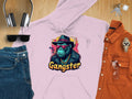 Garment Graphics offers a pink hoodie featuring a gangster gorilla design with a fedora, sunglasses, and chain under Gangster. Displayed on wood with headphones, keys, sunglasses, jeans, a watch, and an orange shirt. Made from medium-heavy fabric for a stylishly cool vibe.