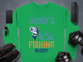 A green Daddys Fishing Buddy sweatshirt by Garment Graphics, featuring a fish graphic, is surrounded by a smartphone, wireless speaker, smartwatch, sneakers, and dumbbells on a dark background—perfect for fishing fans.