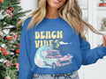 Wearing a blue Garment Graphics sweatshirt featuring Beach Vibes with a classic car, luggage, waves, and palm trees, the person exudes retro vibes beside a beautifully adorned Christmas tree indoors.