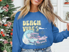 Wearing a blue Garment Graphics sweatshirt featuring Beach Vibes with a classic car, luggage, waves, and palm trees, the person exudes retro vibes beside a beautifully adorned Christmas tree indoors.