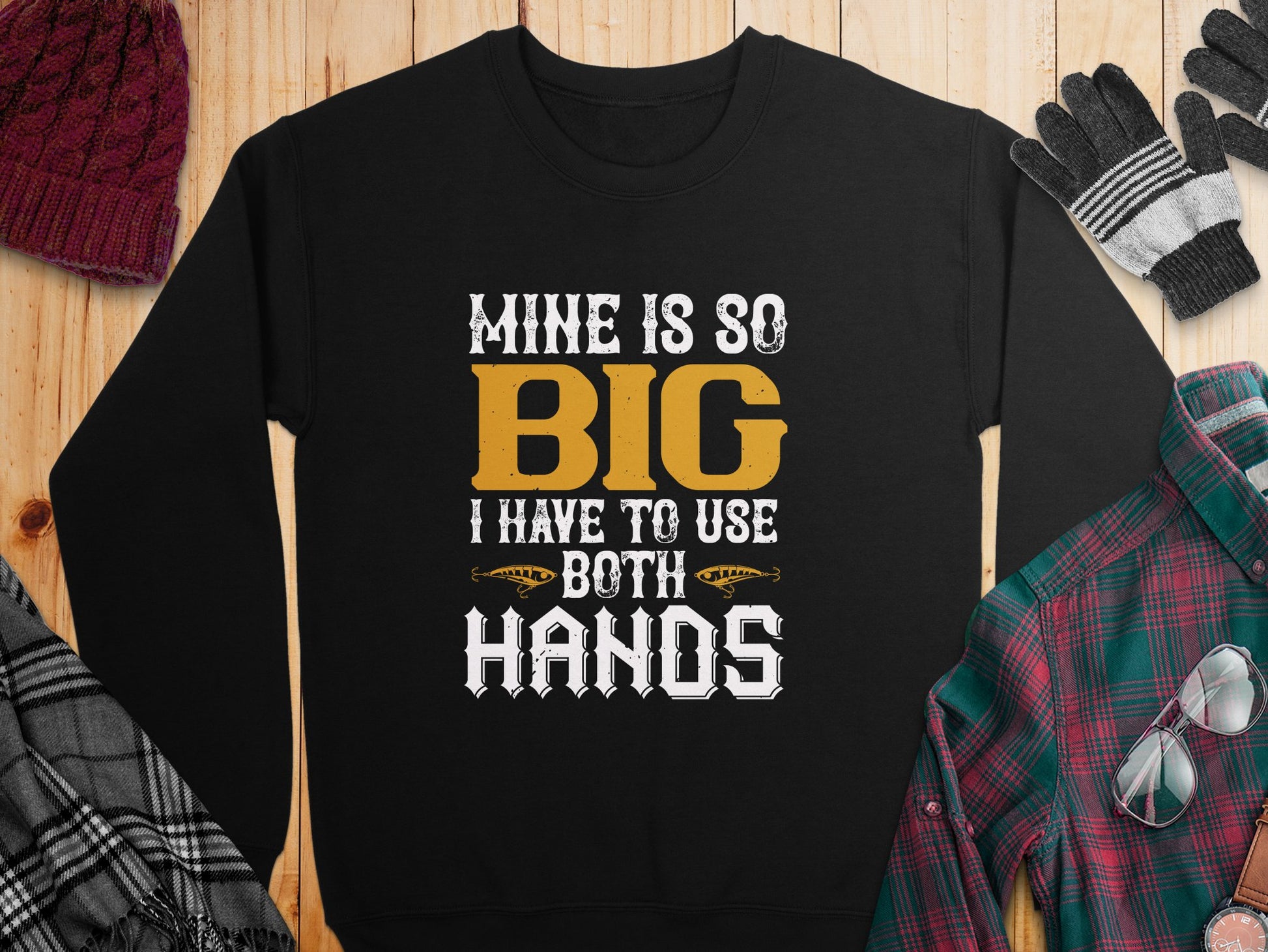 Bold Statement: The Garment Graphics sweatshirt with the text Mine is so big I have to use both hands rests on wood. Paired with a plaid shirt, black and gray gloves, a maroon knit hat, and a black and white scarf, it creates an effortlessly stylish look.