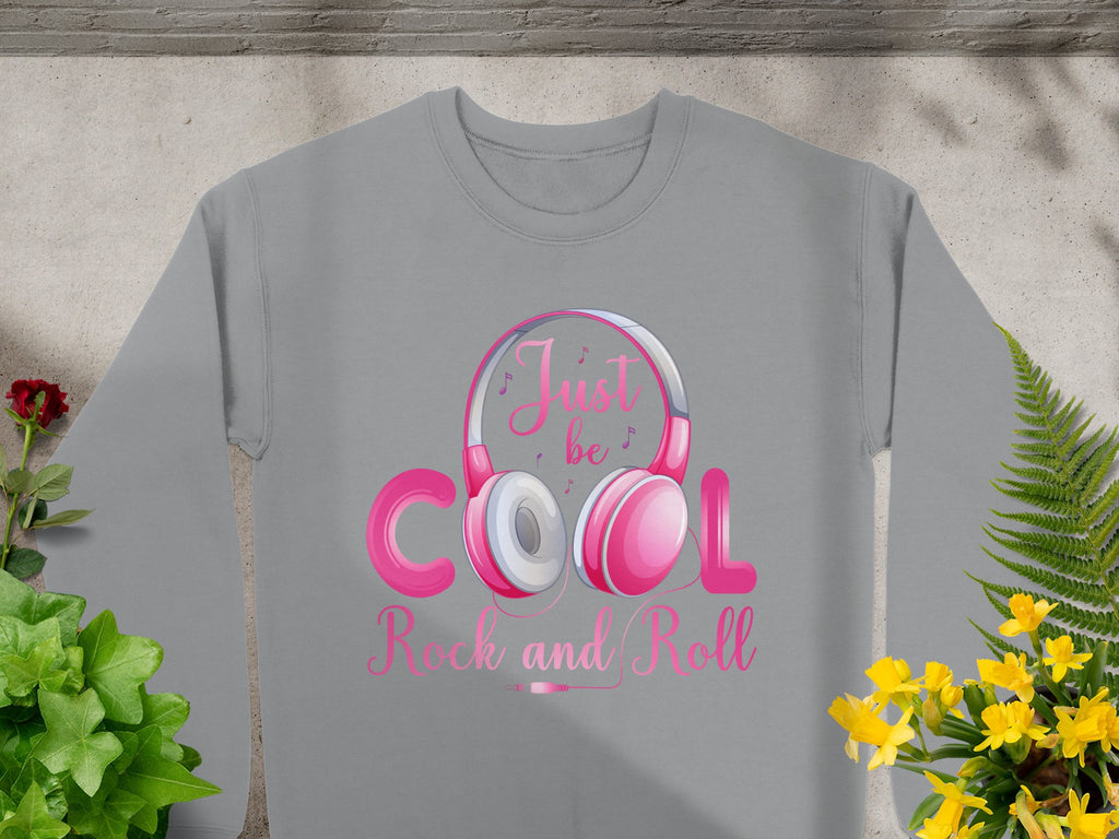 Garment Graphics grey sweatshirt showcases a vibrant pink and white Rock and Roll Headphones graphic with Just be cool, Rock and Roll text. Its adorned with lively yellow, red flowers, and green leaves on a concrete surface—a must-have for music enthusiasts.