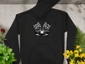 A Garment Graphics black hoodie features a race car and two checkered flags. Laid on concrete, it’s surrounded by green plants, a red flower on the left, and yellow flowers on the right.