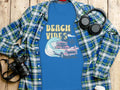 Garment Graphics blue graphic t-shirt showcases Beach Vibes text with a surfing car and palm trees, set on wood. Surrounded by a plaid shirt, headphones, camera, and disposable coffee cup.