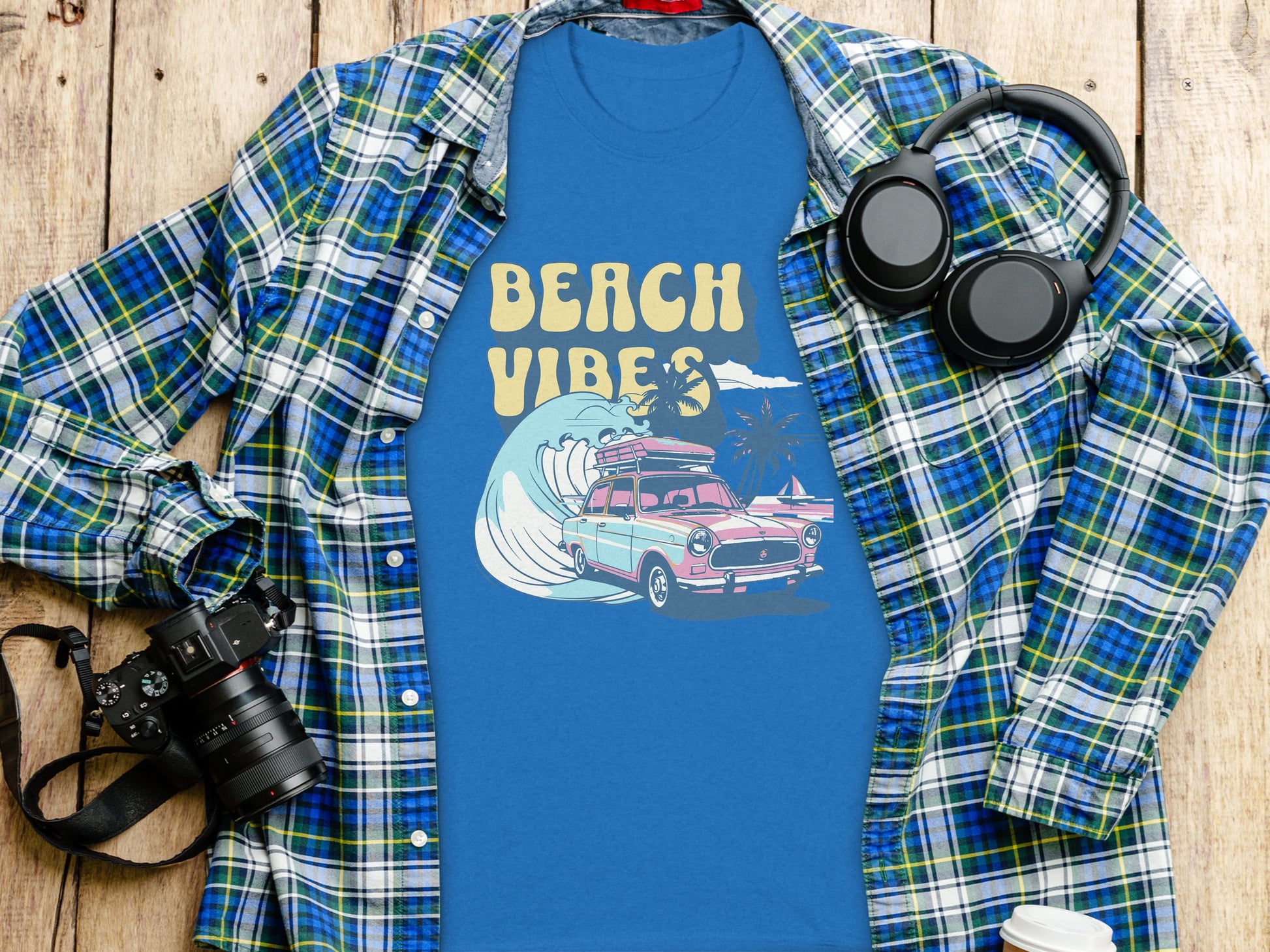 Garment Graphics blue graphic t-shirt showcases Beach Vibes text with a surfing car and palm trees, set on wood. Surrounded by a plaid shirt, headphones, camera, and disposable coffee cup.