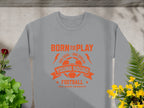 A gray unisex sizing football sweatshirt by Garment Graphics, featuring an orange soccer ball design with text Born to Play, One Goal - One Dream, Young Squads Football, and Kick, Score, Celebrate, lies flat surrounded by flowers and ferns.