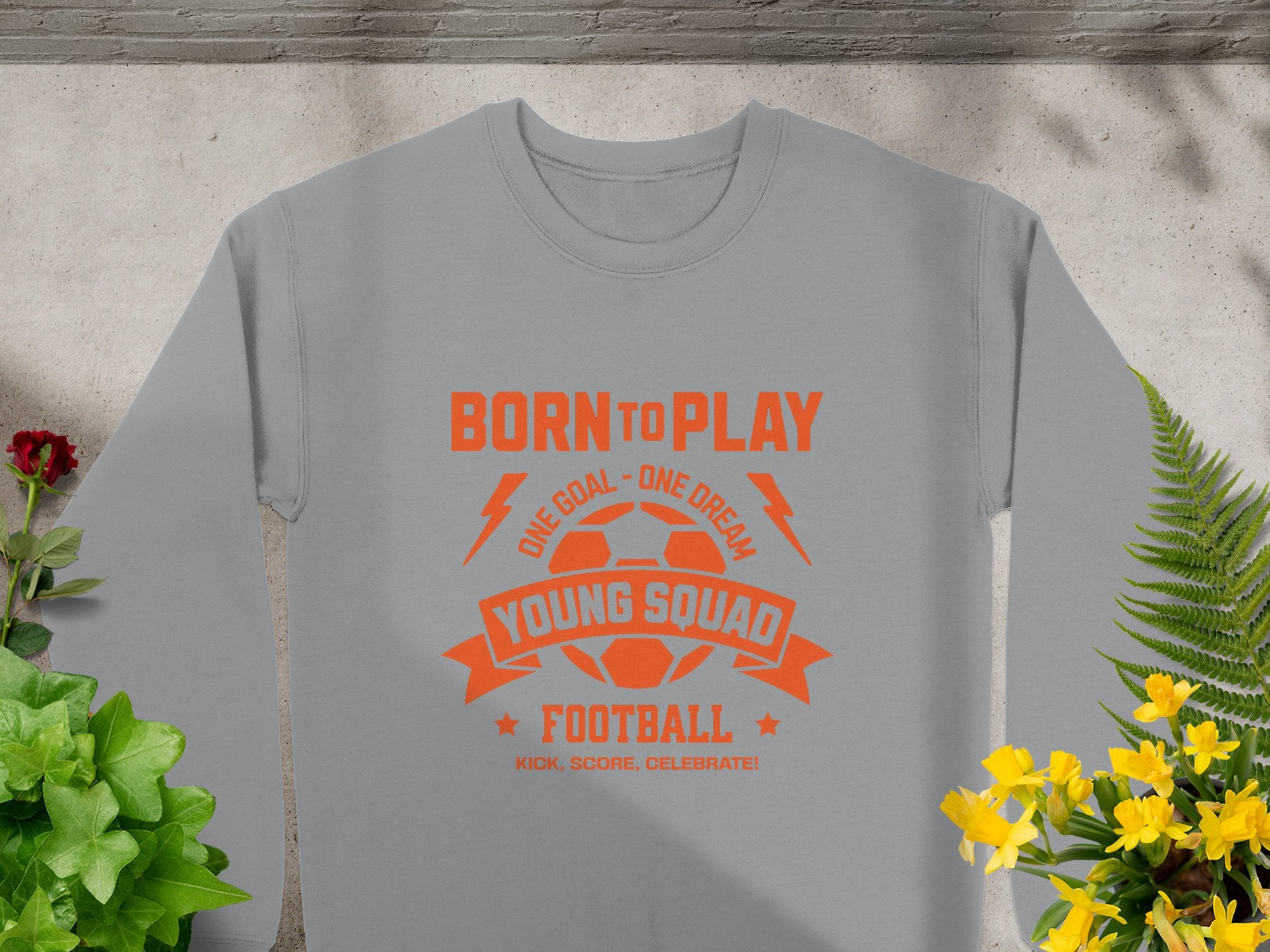 A gray unisex sizing football sweatshirt by Garment Graphics, featuring an orange soccer ball design with text Born to Play, One Goal - One Dream, Young Squads Football, and Kick, Score, Celebrate, lies flat surrounded by flowers and ferns.