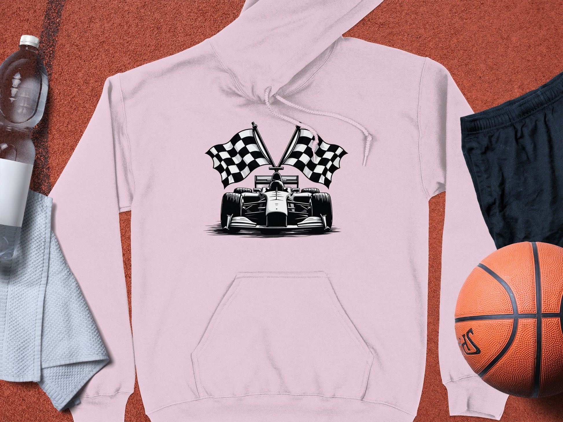 The Garment Graphics hoodie features a light pink hue with a race car and racing flags design, ideal for motorsport fans. Its set among a clear water bottle, light gray towel, black shorts, and orange basketball on a textured reddish-brown background.