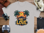Garment Graphics offers a light gray T-shirt ideal for beach lovers, showcasing a tropical sunset print with palm trees, a plane, and “ONE LOVE” boldly displayed. Styled on wood and paired with essentials like a camera, binoculars, wallet, and small bag.