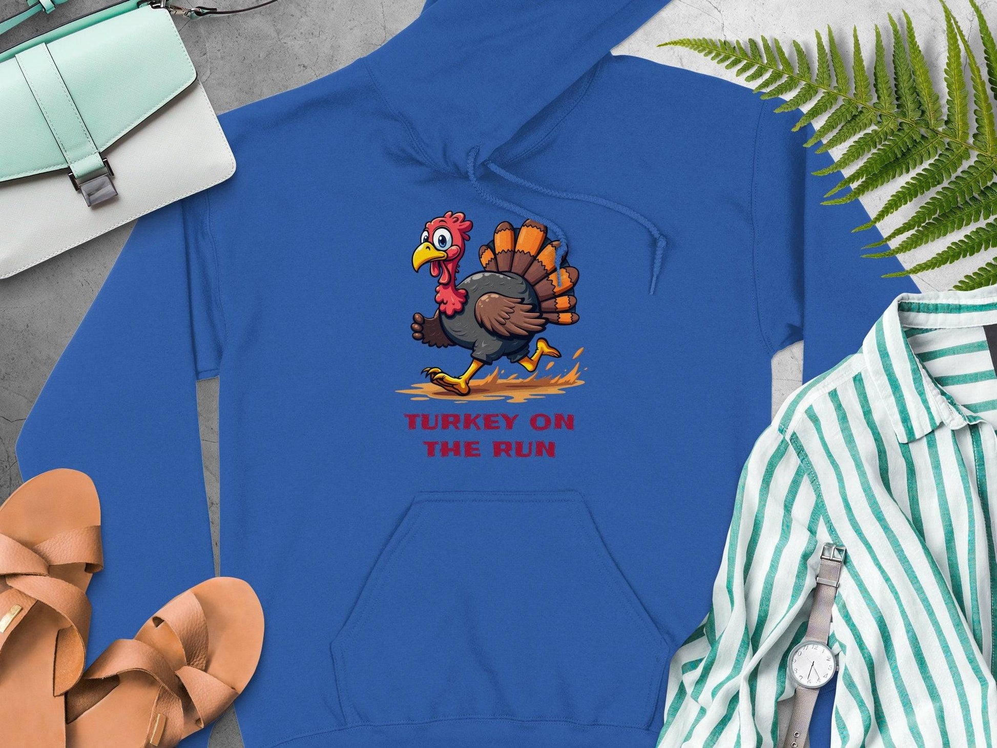 This blue hoodie by Garment Graphics features a cartoon turkey with Turkey on the Run printed beneath. Its displayed with sandals, a striped shirt, wristwatch, and light blue bag on a flat surface, accented by fern leaves for a fresh touch.