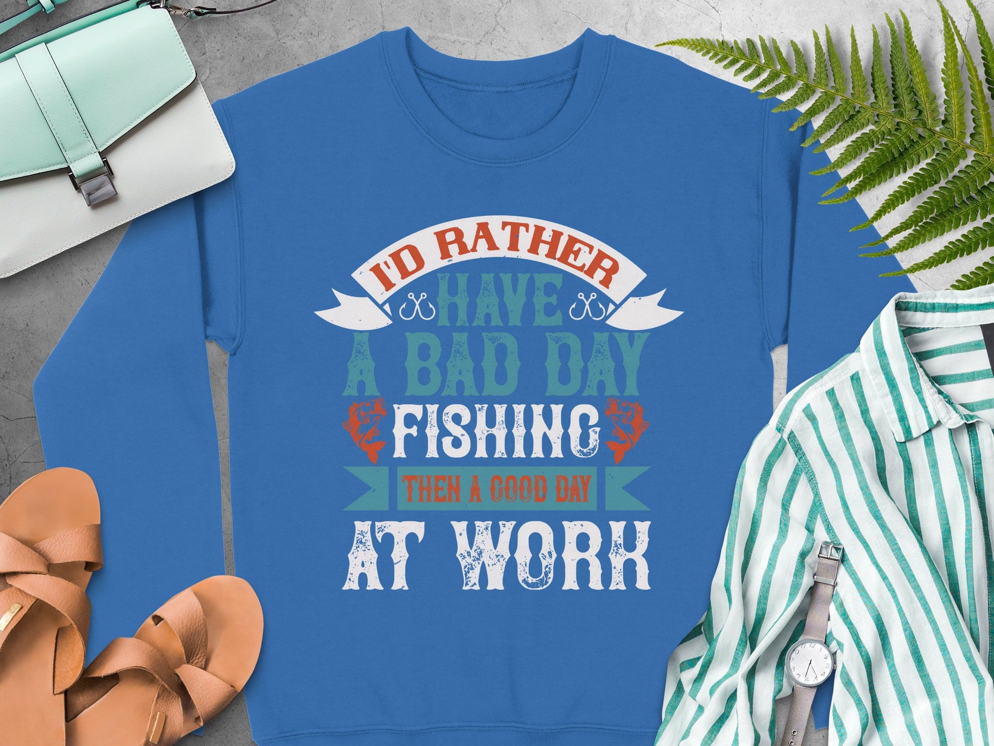 A blue unisex fishing sweatshirt from Garment Graphics, with Id rather have a bad day fishing than a good day at work, is set on concrete. Around it are a green purse, tan sandals, a striped shirt, and some fern leaves.