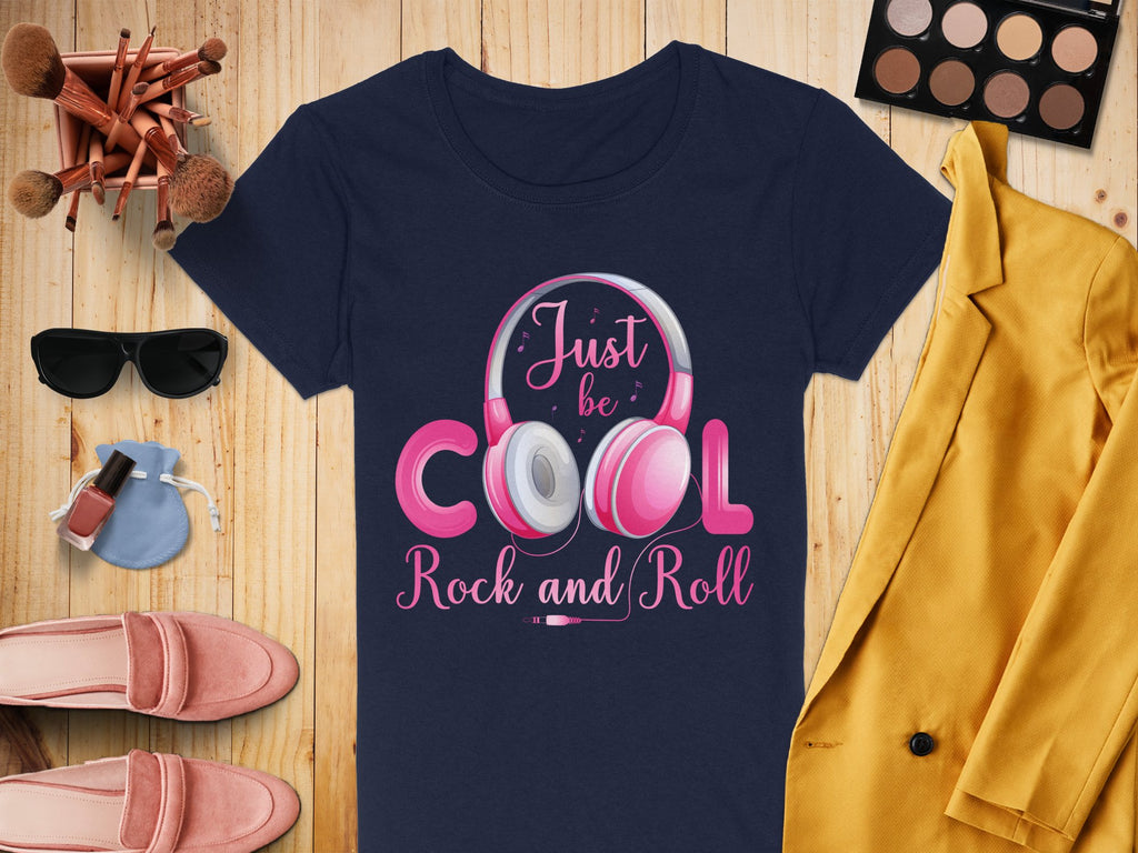Flat lay for music lovers showcasing a Garment Graphics navy headphones T-shirt with Just be Cool Rock and Roll design, surrounded by a yellow coat, makeup palette, brushes, sunglasses, pink shoes, nail polish, and a small blue pouch on a wooden surface.