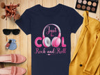 Flat lay for music lovers showcasing a Garment Graphics navy headphones T-shirt with Just be Cool Rock and Roll design, surrounded by a yellow coat, makeup palette, brushes, sunglasses, pink shoes, nail polish, and a small blue pouch on a wooden surface.