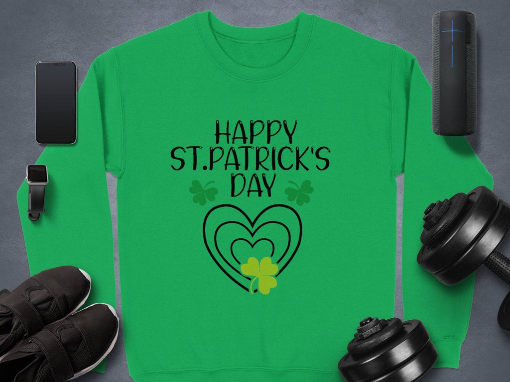 The Garment Graphics festive green sweatshirt features Happy St. Patricks Day text with bold black clover designs, surrounded by a smartphone, smartwatch, wireless speaker, sneakers, and dumbbells on a gray surface.