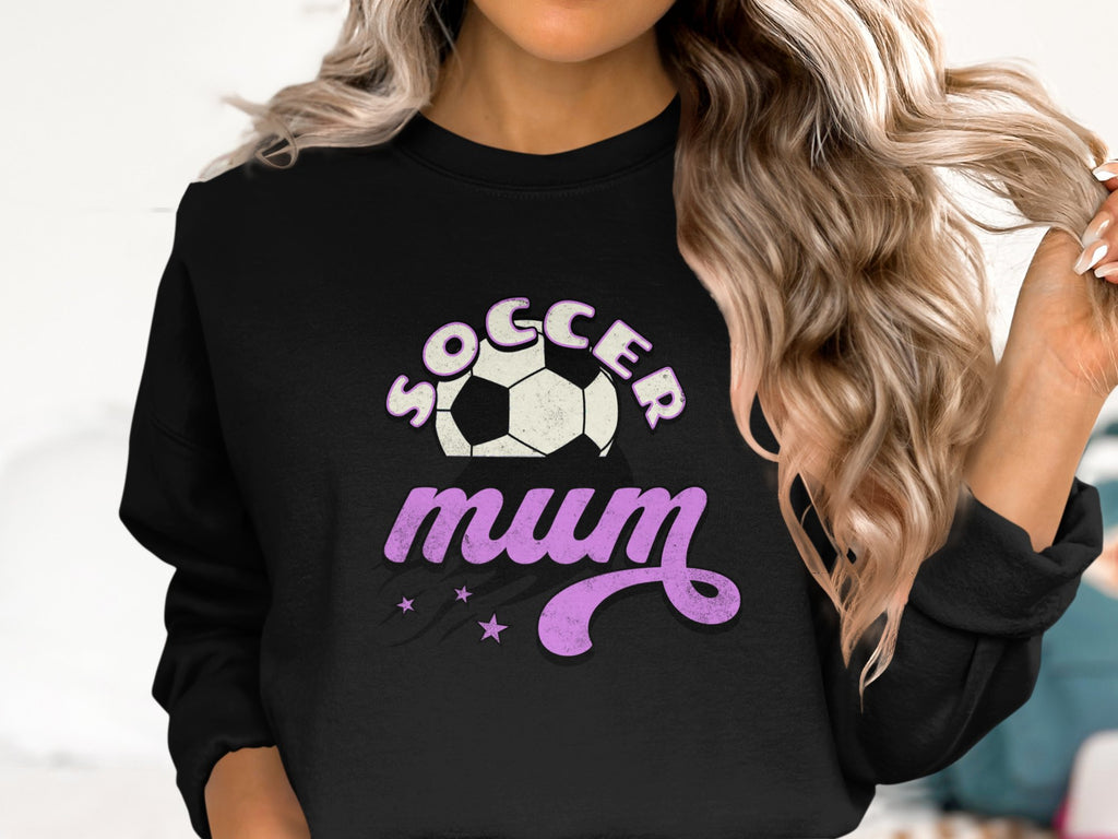 A person with long, wavy blonde hair wears a black Garment Graphics sweatshirt featuring Soccer Mum in white and purple text, accompanied by a soccer ball graphic and vintage-style purple stars.
