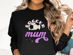 A person with long, wavy blonde hair wears a black Garment Graphics sweatshirt featuring Soccer Mum in white and purple text, accompanied by a soccer ball graphic and vintage-style purple stars.