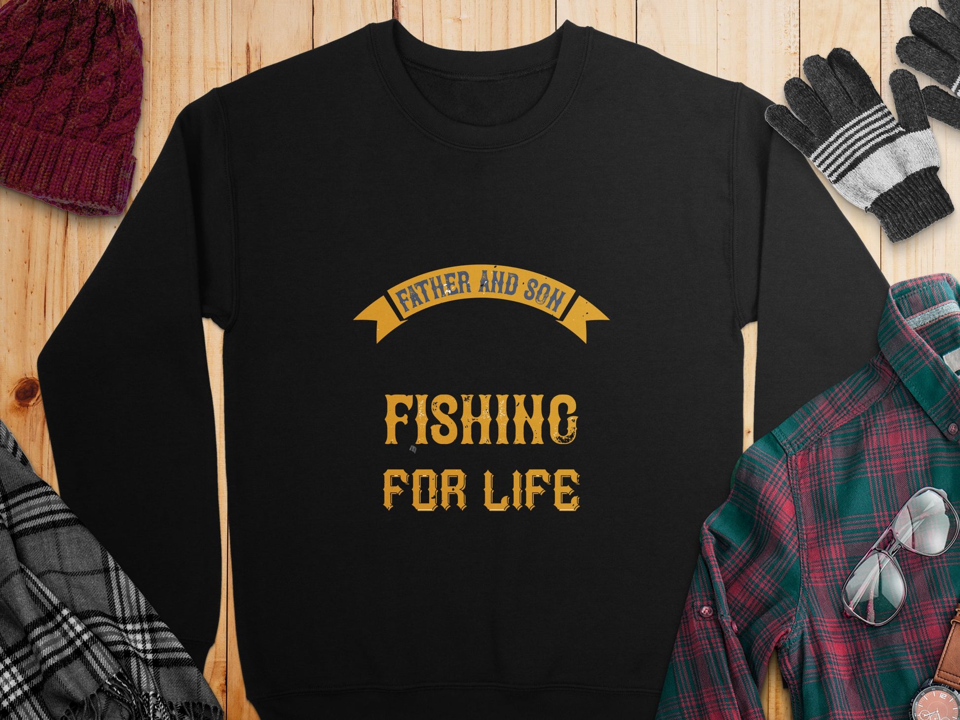 A black graphic sweatshirt from Garment Graphics lays flat on a wooden surface, featuring the slogan Father and Son Fishing for Life in bold gold letters. Its surrounded by a red knit hat, gray gloves, a black and white scarf, and a green plaid shirt.
.