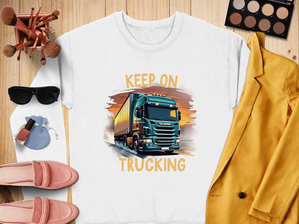 A Garment Graphics white graphic sweatshirt featuring a truck and Keep on Trucking rests on a wooden surface, ideal for truck fans. Its styled with pink shoes, lipstick, sunglasses, makeup brushes, a yellow jacket, and an eyeshadow palette.