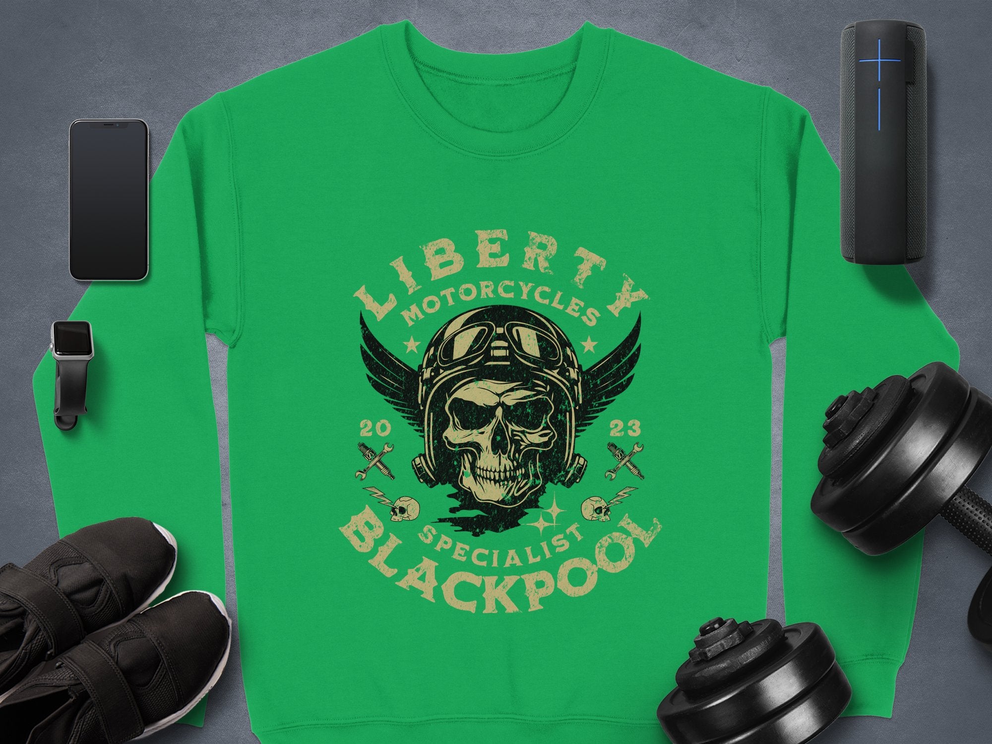A Garment Graphics green sweatshirt features a black skull with goggles and helmet, surrounded by Liberty Motorcycles Blackpool Specialist 2023 and crossed wrenches. Complete your biker style with items like a phone, speaker, dumbbells, shoes, and smartwatch nearby.