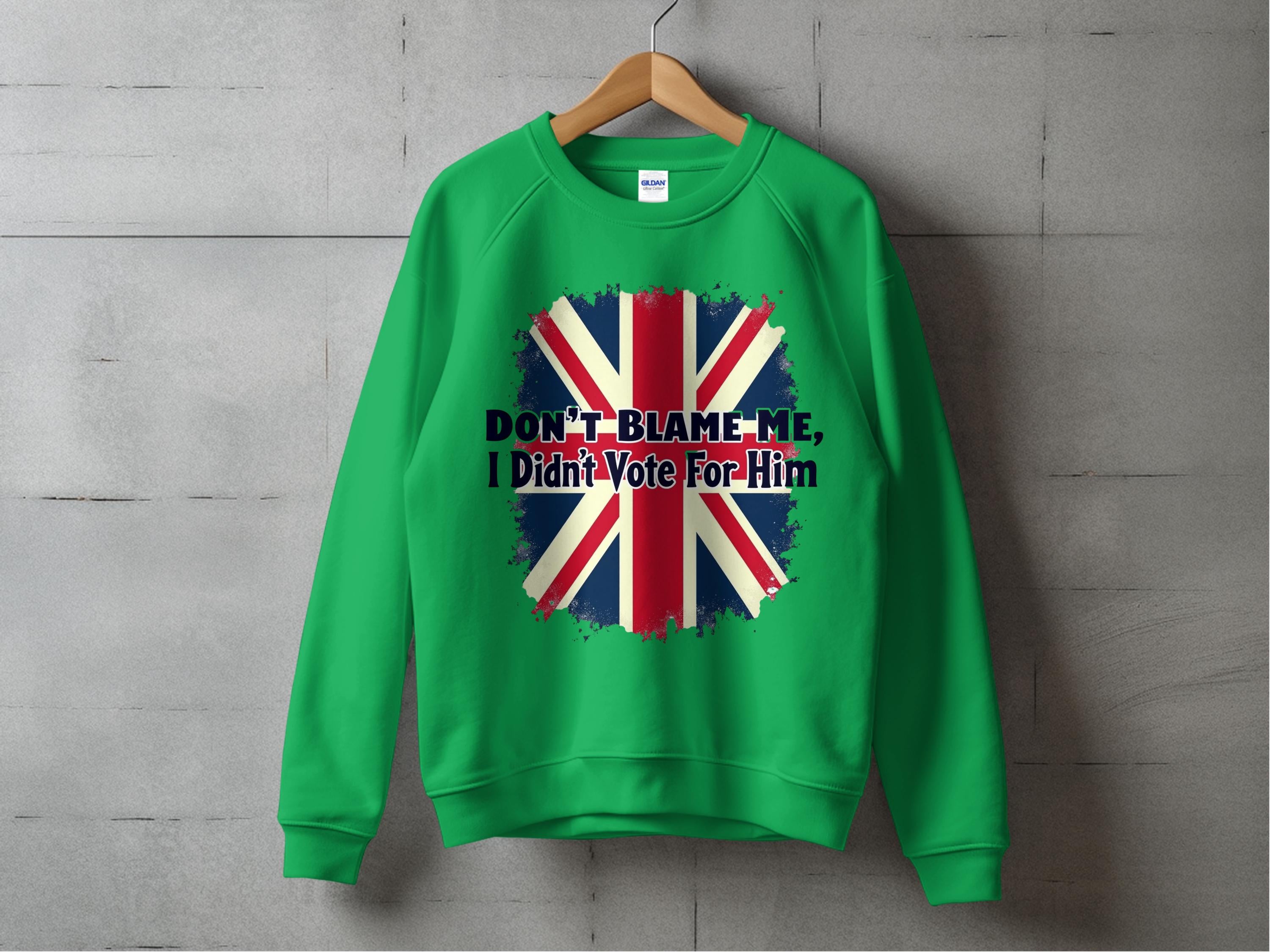 A comfortable green sweatshirt by Garment Graphics hangs on the wall, featuring a bold design with a distressed Union Jack and the striking text DONT BLAME ME, I DIDNT VOTE FOR HIM printed over the flag.