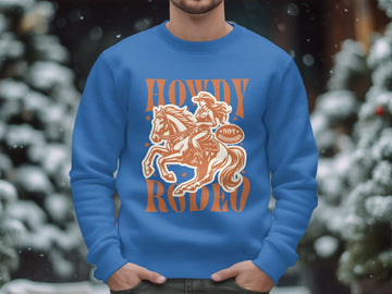 A person is wearing a Garment Graphics unisex graphic print sweatshirt featuring an illustrated cowboy on horseback with the words Howdy Rodeo and Notorious. Snow-covered trees in the blurred background enhance its charm.