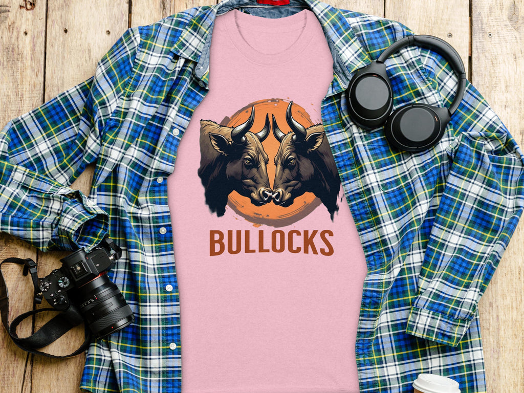 A pink Garment Graphics Pintable T-Shirt featuring two bull heads above BULLOCKS is displayed on wood, surrounded by a blue and green plaid shirt, a camera, headphones, and a white paper cup. Enjoy matching family styles in various sizes.