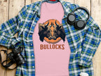 A pink Garment Graphics Pintable T-Shirt featuring two bull heads above BULLOCKS is displayed on wood, surrounded by a blue and green plaid shirt, a camera, headphones, and a white paper cup. Enjoy matching family styles in various sizes.