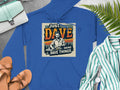 Garment Graphics blue graphic hoodie features Just Dave Doing Dave Things with a mans illustration. Ideal for casual outings, it offers medium-heavy fabric for style and comfort. Pair it with sandals, a light blue handbag, and a striped shirt against fern leaves on a gray background.