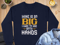 A Garment Graphics navy blue sweatshirt with Mine is so big I have to use both hands in yellow and white letters lies on a wooden surface, paired with a blue denim jacket and a steaming latte nearby.