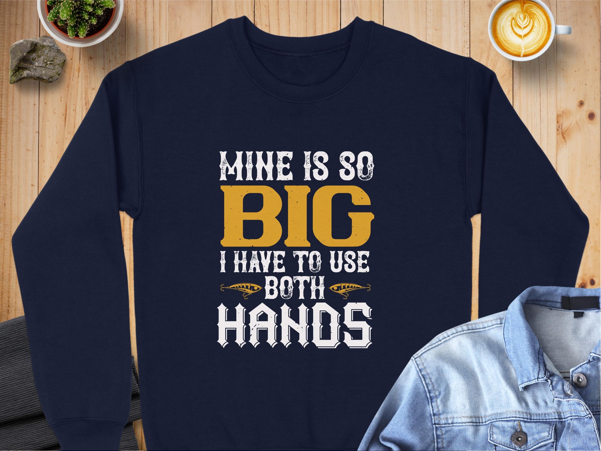 A Garment Graphics navy blue sweatshirt with Mine is so big I have to use both hands in yellow and white letters lies on a wooden surface, paired with a blue denim jacket and a steaming latte nearby.