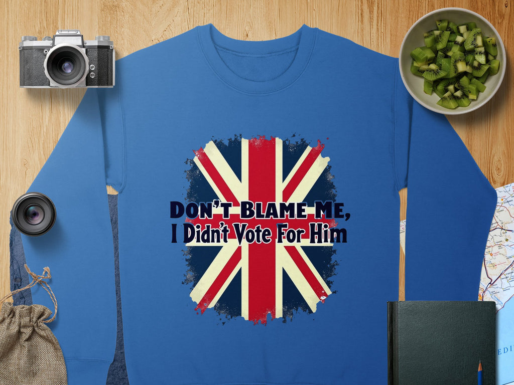 A blue unisex sweatshirt from Garment Graphics features a Union Jack and the text Dont Blame Me, I Didnt Vote For Him. Its presented on a wooden surface surrounded by a camera, lenses, chopped kiwi, a map, and a small drawstring bag.