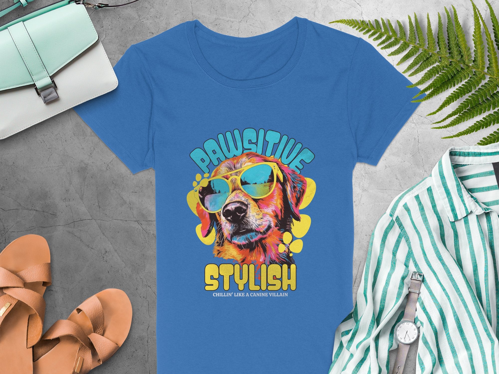 Garment Graphics blue dog T-shirt showcases a fun graphic of a dog in sunglasses and a hat with Pawsitive above and Stylish below. Ideal for adding flair, it complements sandals, purse, watch, striped shirt, and ferns on gray surfaces. Available in womens sizes.