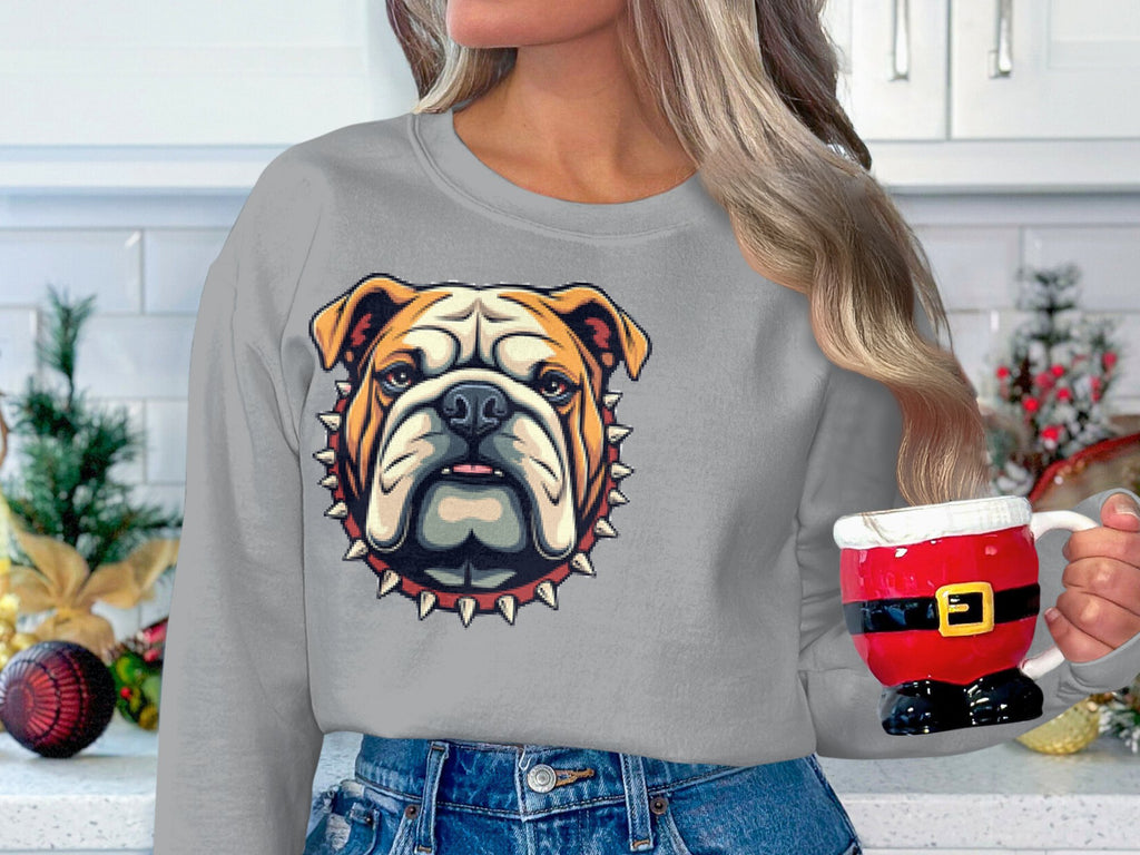 In a festive kitchen decorated with pine and ornaments, a long-haired person holds a Santa mug, wearing Garment Graphics unisex gray sweatshirt featuring a bold bulldog design with spikes—perfect for the holiday spirit.