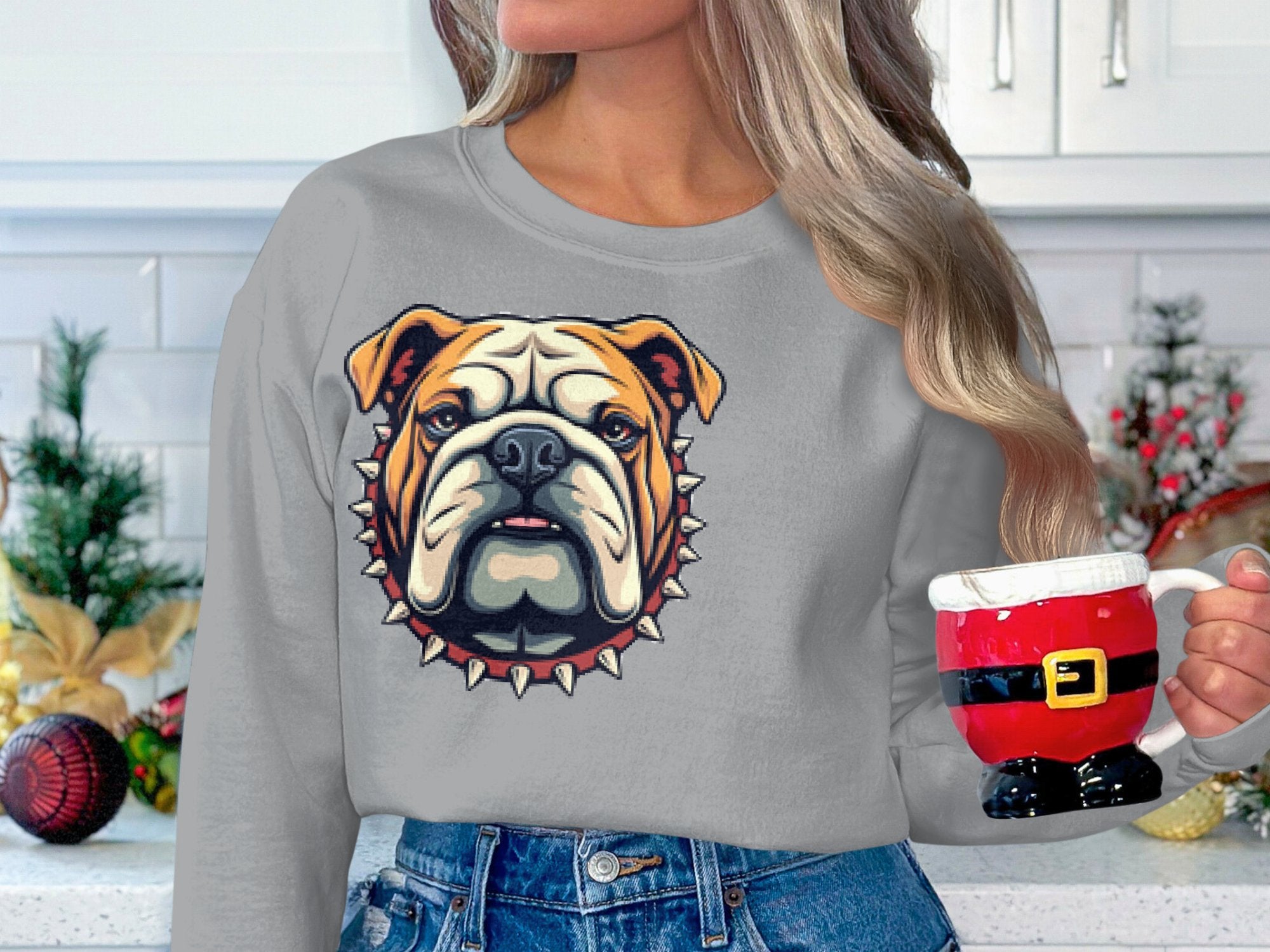 In a festive kitchen decorated with pine and ornaments, a long-haired person holds a Santa mug, wearing Garment Graphics unisex gray sweatshirt featuring a bold bulldog design with spikes—perfect for the holiday spirit.