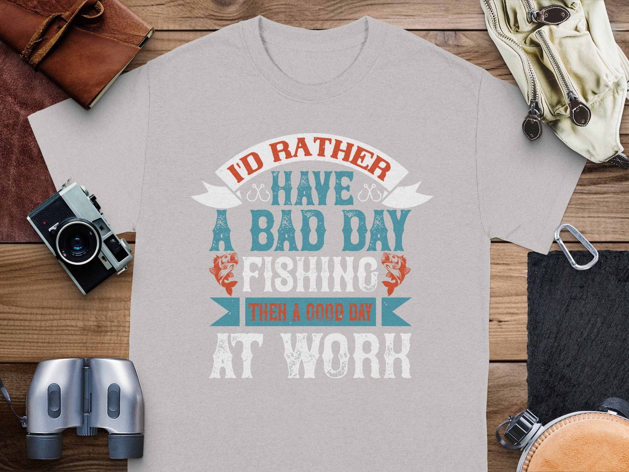 A light gray fishing T-shirt by Garment Graphics showcases the message: Id rather have a bad day fishing than a good day at work. This ideal gift for anglers is set on a wooden surface with items like a leather wallet, vintage camera, binoculars, and a belt nearby.