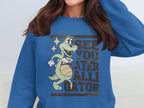 A woman with long brown hair wears a blue Garment Graphics sweatshirt featuring a playful alligator graphic holding a ball and the text See You Later Alligator, making it an ideal standout for casual outings.