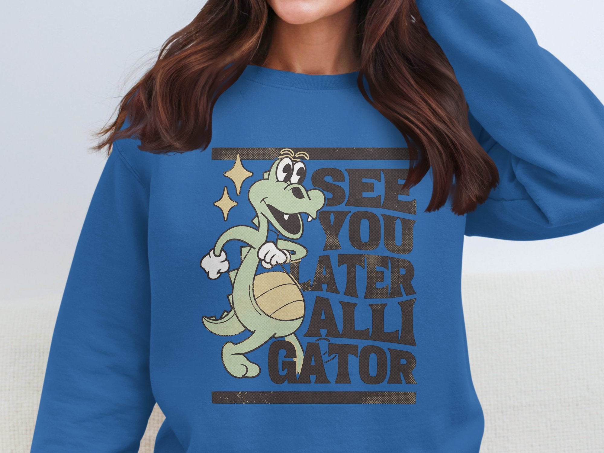 A woman with long brown hair wears a blue Garment Graphics sweatshirt featuring a playful alligator graphic holding a ball and the text See You Later Alligator, making it an ideal standout for casual outings.