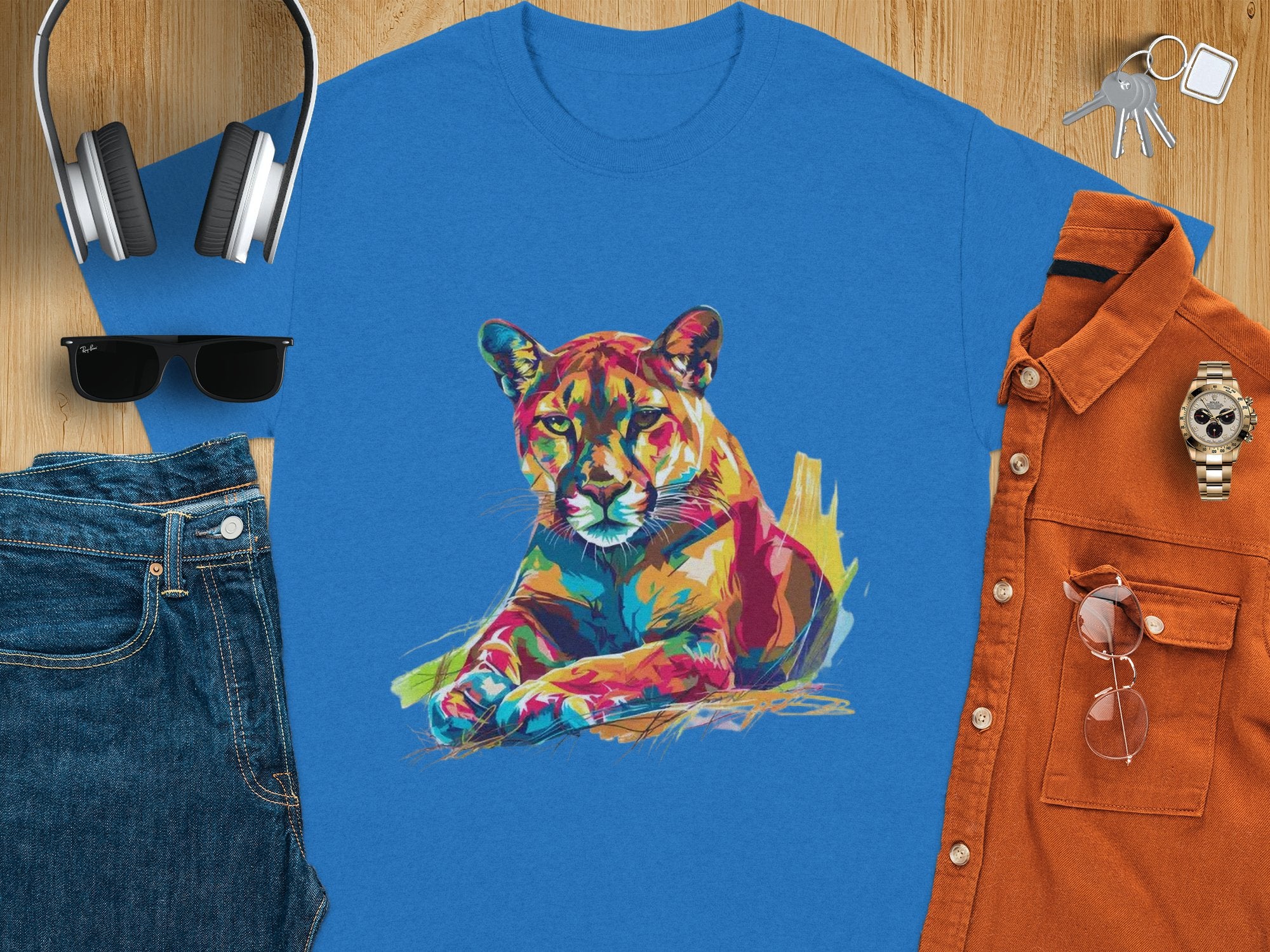 A Garment Graphics blue T-shirt with vibrant cougar artwork rests on a wooden surface, surrounded by jeans, sunglasses, headphones, a watch, keys, and an orange button-up shirt.