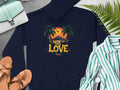 A Garment Graphics navy blue hoodie displays a tropical sunset, palm trees, and One Love. It lies beside sandals, a striped shirt, watch, purse, and ferns on gray.