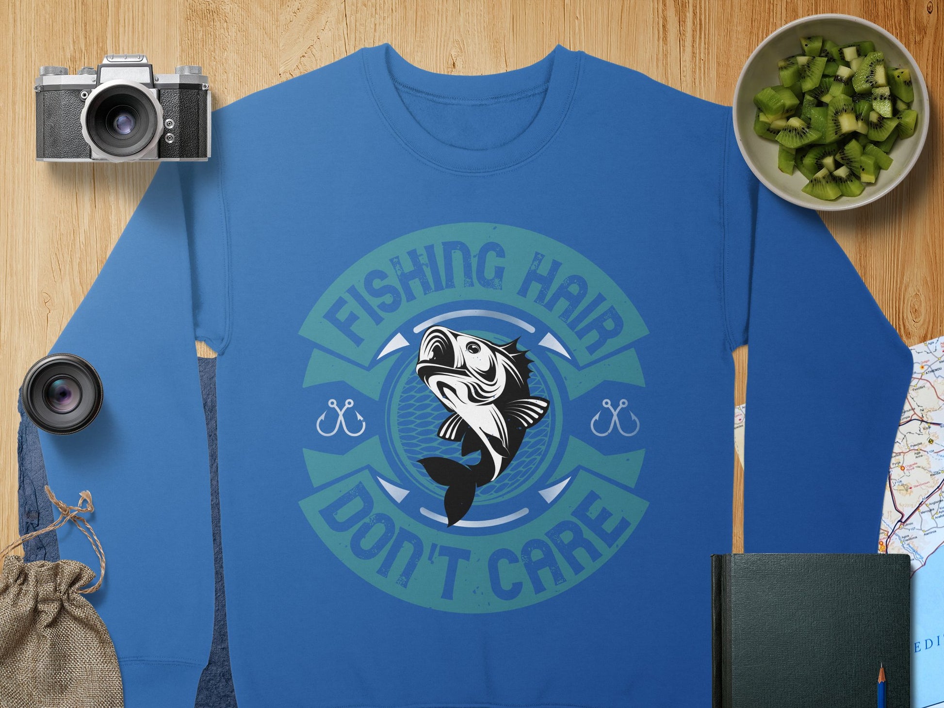 A Garment Graphics blue fishing sweatshirt with a vibrant fish design and the text Fishing Hair Don’t Care is displayed on wood. Nearby are fishing essentials: a camera, lens, small fruit bowl, map, green book, and drawstring bag.