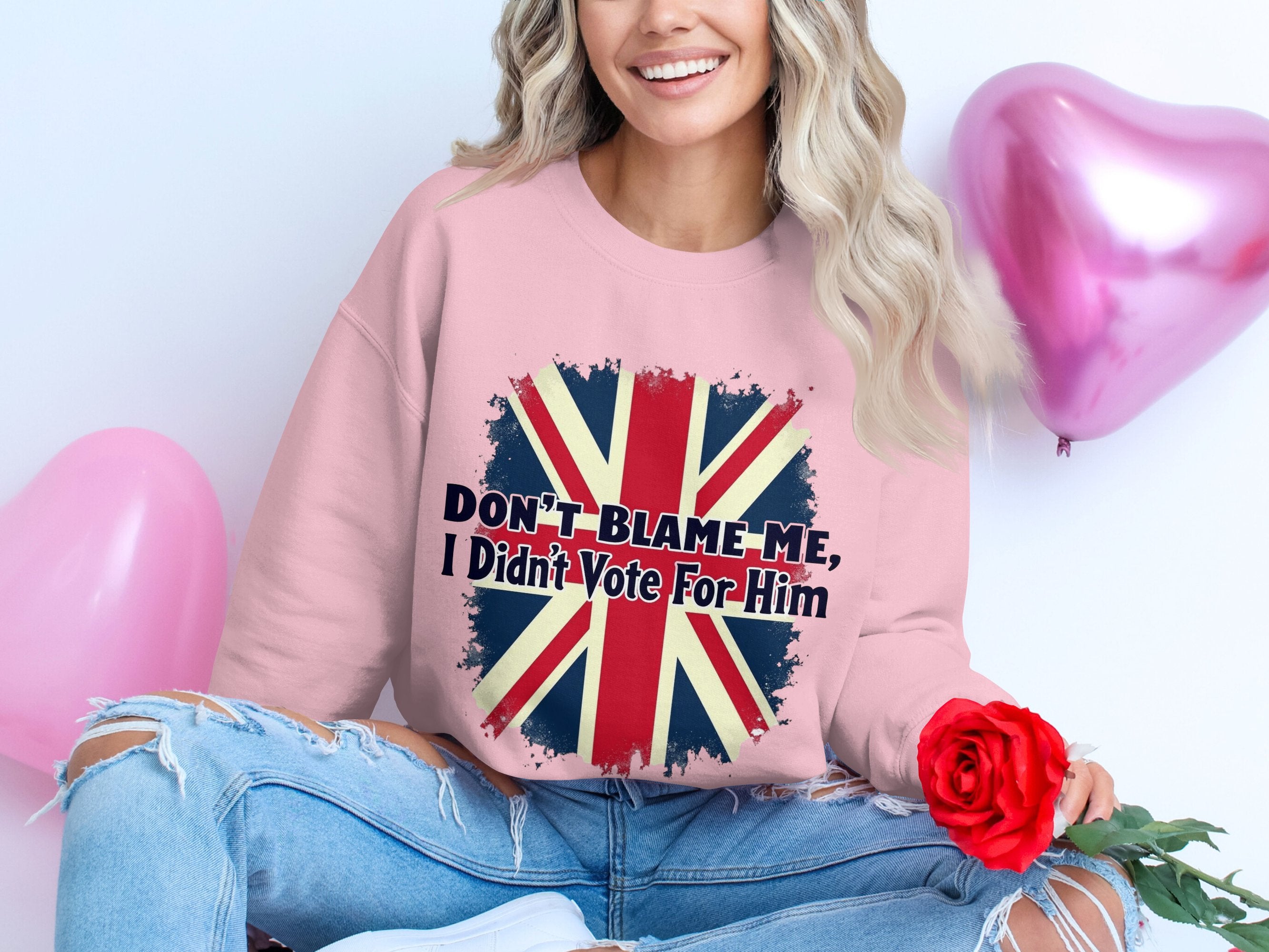 A person wears a Garment Graphics pink sweatshirt with the Union Jack and Dont Blame Me, I Didnt Vote For Him text in trendy unisex sizing. They hold a red rose beside pink heart-shaped balloons, smiling with wavy hair in stylishly ripped jeans at the knees.