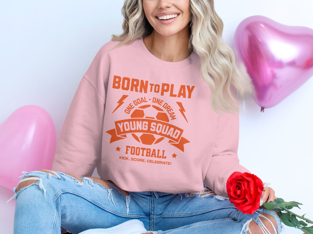 A person in a pink Garment Graphics football sweatshirt with the text Born to Play, One Goal - One Dream, Young Squad, Football. holds a red rose while sitting near shiny pink heart balloons. Embracing unisex sizing, this young squad expresses their passion stylishly.