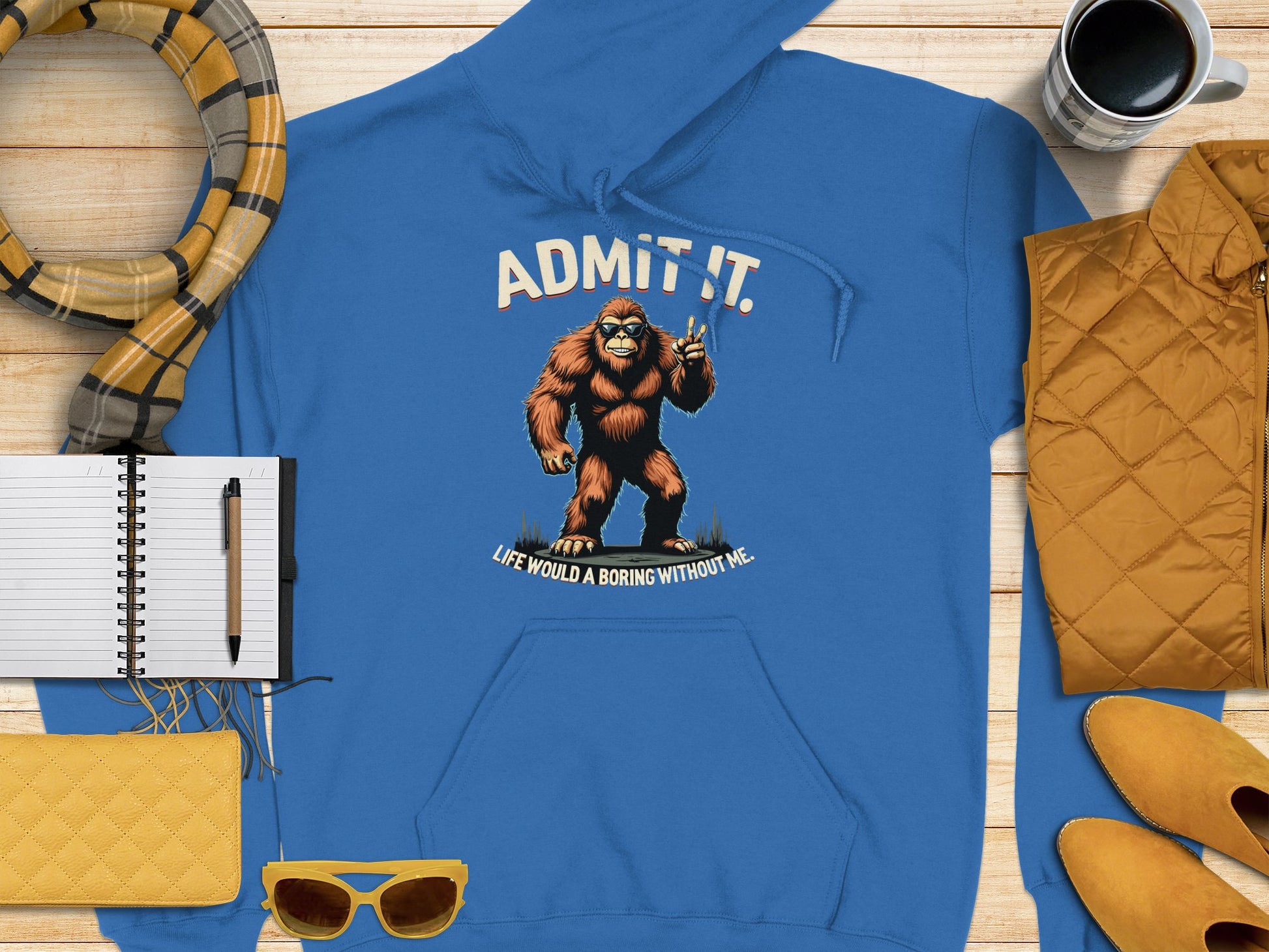 A Garment Graphics blue classic fit hoodie featuring Bigfoot and the phrase Admit it. Life would be boring without me, is laid on wood, surrounded by a plaid scarf, notebook, pen, coffee cup, yellow purse, sunglasses, yellow vest, and shoes—a display exuding cool character.