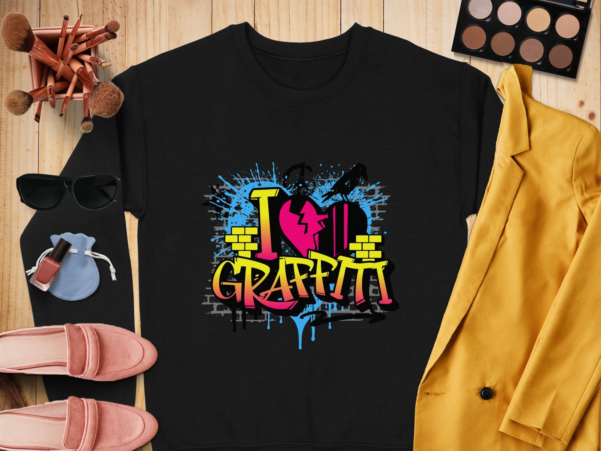 A black sweatshirt from Garment Graphics featuring I love graffiti in vibrant design rests on wood, encircled by sunglasses, makeup brushes, a palette, a yellow jacket, pink shoes, and a blue pouch—ideal for an urban style enthusiast.