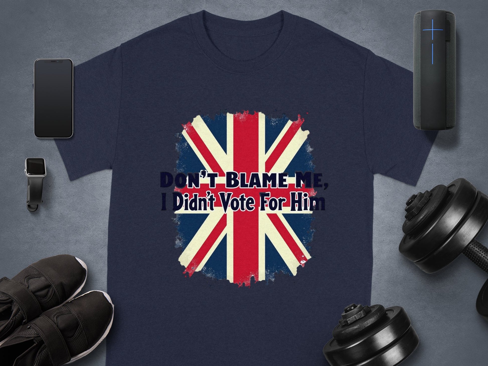 A navy blue classic-fit T-shirt by Garment Graphics features a Union Jack and DONT BLAME ME, I Didnt Vote For Him. It is accompanied by a smartwatch, wireless earphones, smart speaker, dumbbells, and black sneakers.