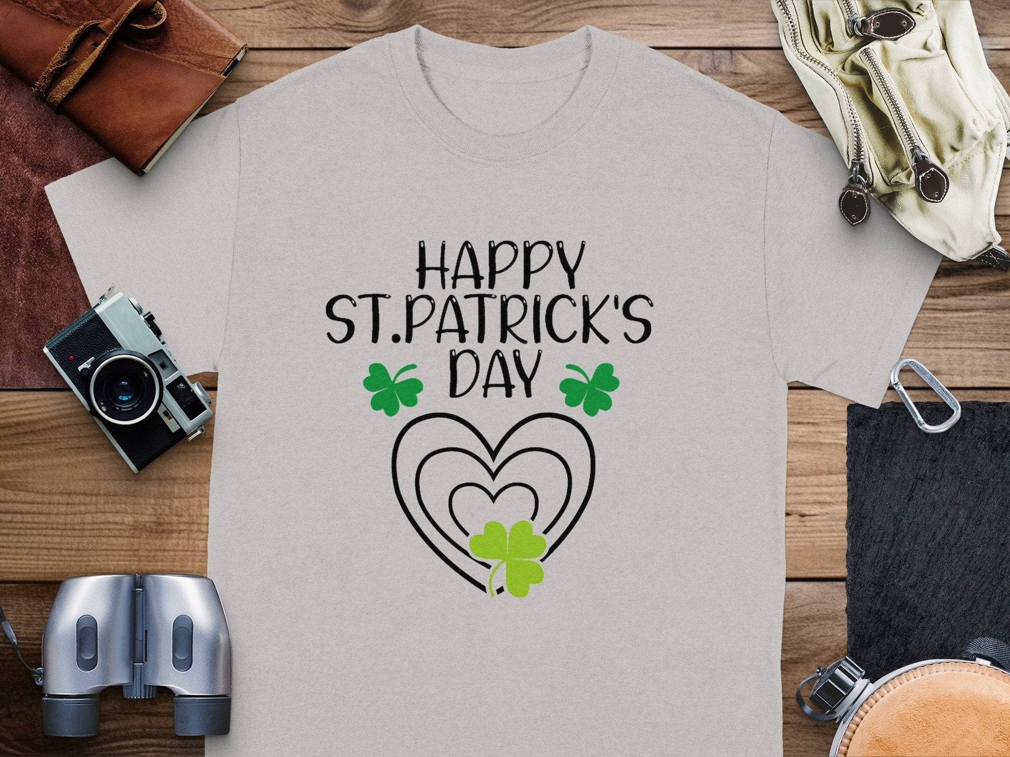 The Garment Graphics gray graphic T-shirt, adorned with Happy St. Patricks Day in black, features green shamrocks and a heart design. It exudes festive flair as it rests on a wooden surface surrounded by a camera, leather items, binoculars, and gloves.