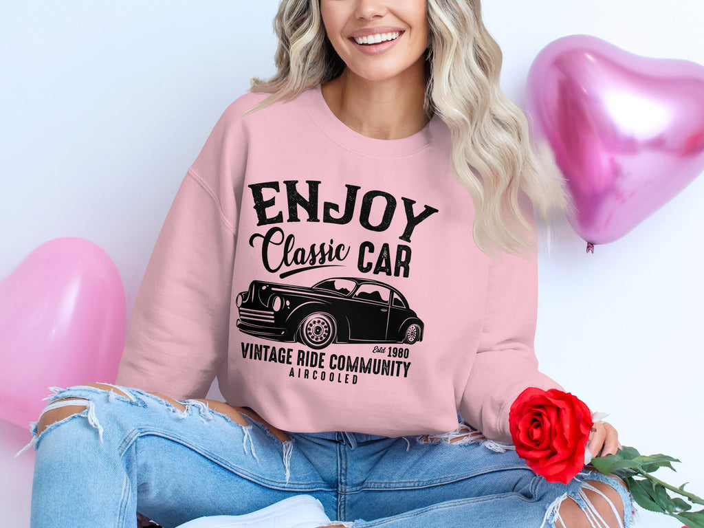 A woman in a pink Vintage Ride sweatshirt by Garment Graphics, featuring Enjoy Classic Car, Vintage Ride Community, Aircooled Est. 1980, holds a red rose. She sits on the floor in ripped jeans beside pink heart-shaped balloons, exuding classic car charm.