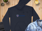 A trendy black hoodie from Garment Graphics, featuring the iconic Scania logo, is laid flat on a wooden surface, surrounded by a denim jacket, a latte with art, a potted plant, and a stone.