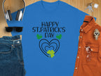 A blue graphic T-shirt from Garment Graphics features Happy St. Patricks Day in black, surrounded by shamrocks and hearts. Its showcased with headphones, keys, a brown shirt, watch, sunglasses, and denim jeans on a wooden surface for a festive look.