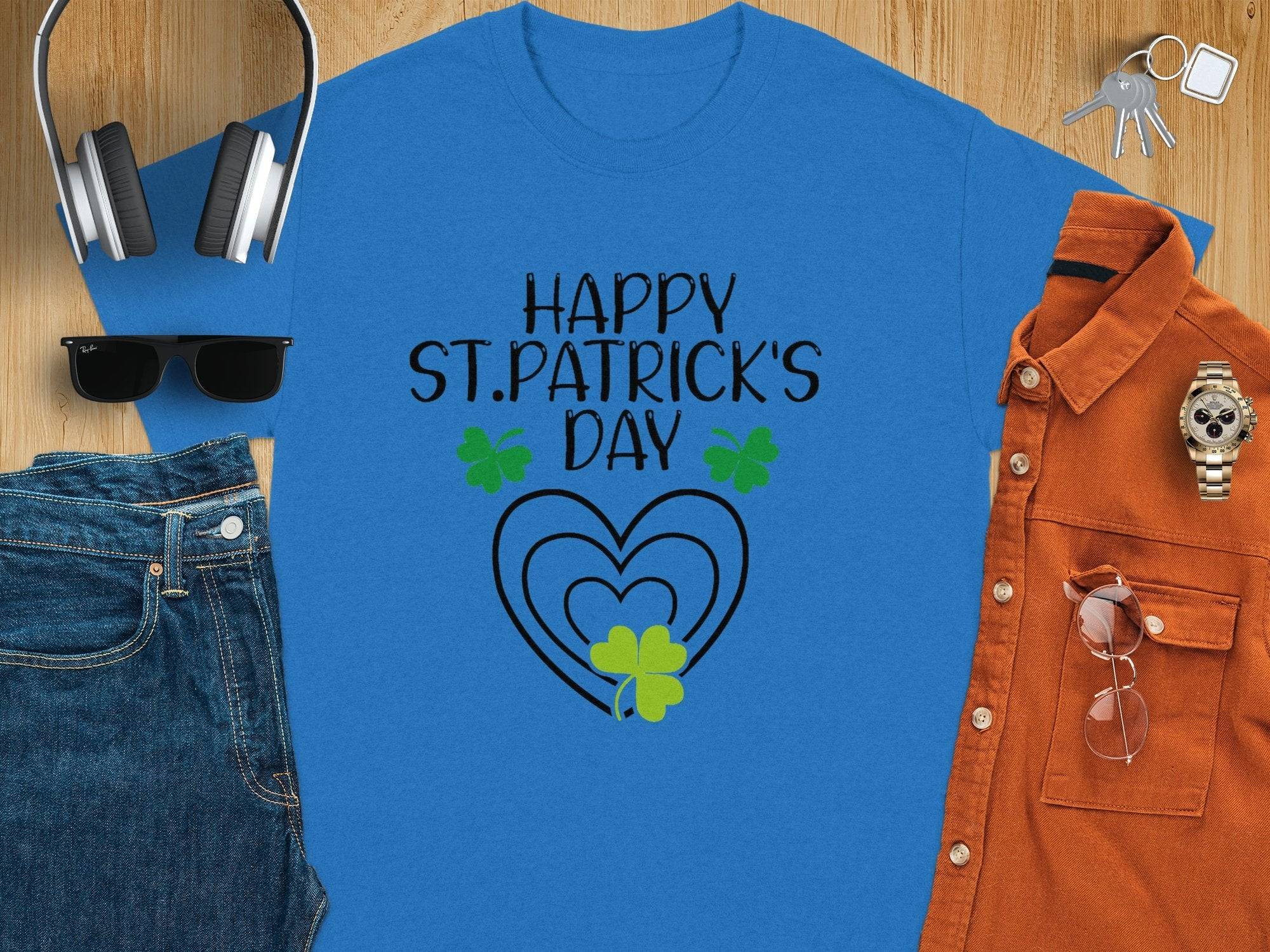 A blue graphic T-shirt from Garment Graphics features Happy St. Patricks Day in black, surrounded by shamrocks and hearts. Its showcased with headphones, keys, a brown shirt, watch, sunglasses, and denim jeans on a wooden surface for a festive look.